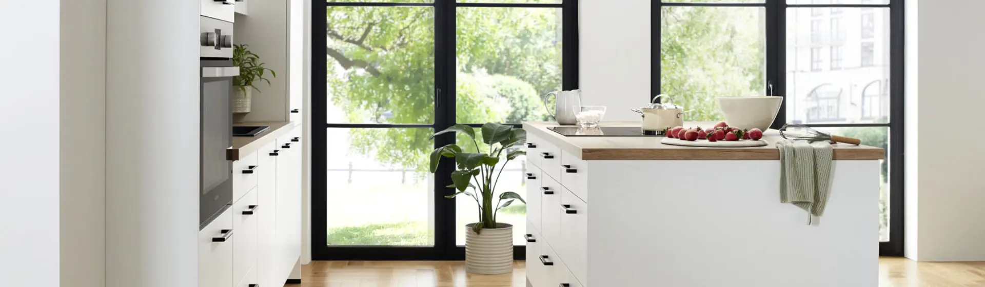 Helle, moderne Küche mit weißen Schränken und einer Holzinsel. Große Fenster geben den Blick ins Grüne frei Helle, moderne Küche mit weißen Schränken und einer Holzinsel. Große Fenster geben den Blick ins Grüne frei