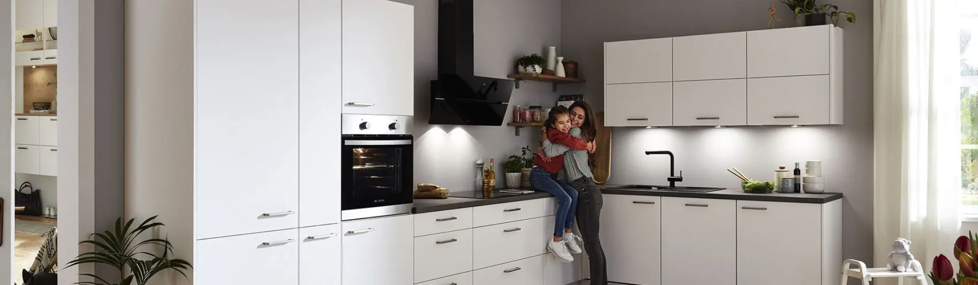 Moderne Küche mit weißen Schränken, schwarzen Arbeitsplatten und Edelstahlofen. Eine Frau und ein Kind sitzen auf der Theke und umarmen sich freudig Moderne Küche mit weißen Schränken, schwarzen Arbeitsplatten und Edelstahlofen. Eine Frau und ein Kind sitzen auf der Theke und umarmen sich freudig